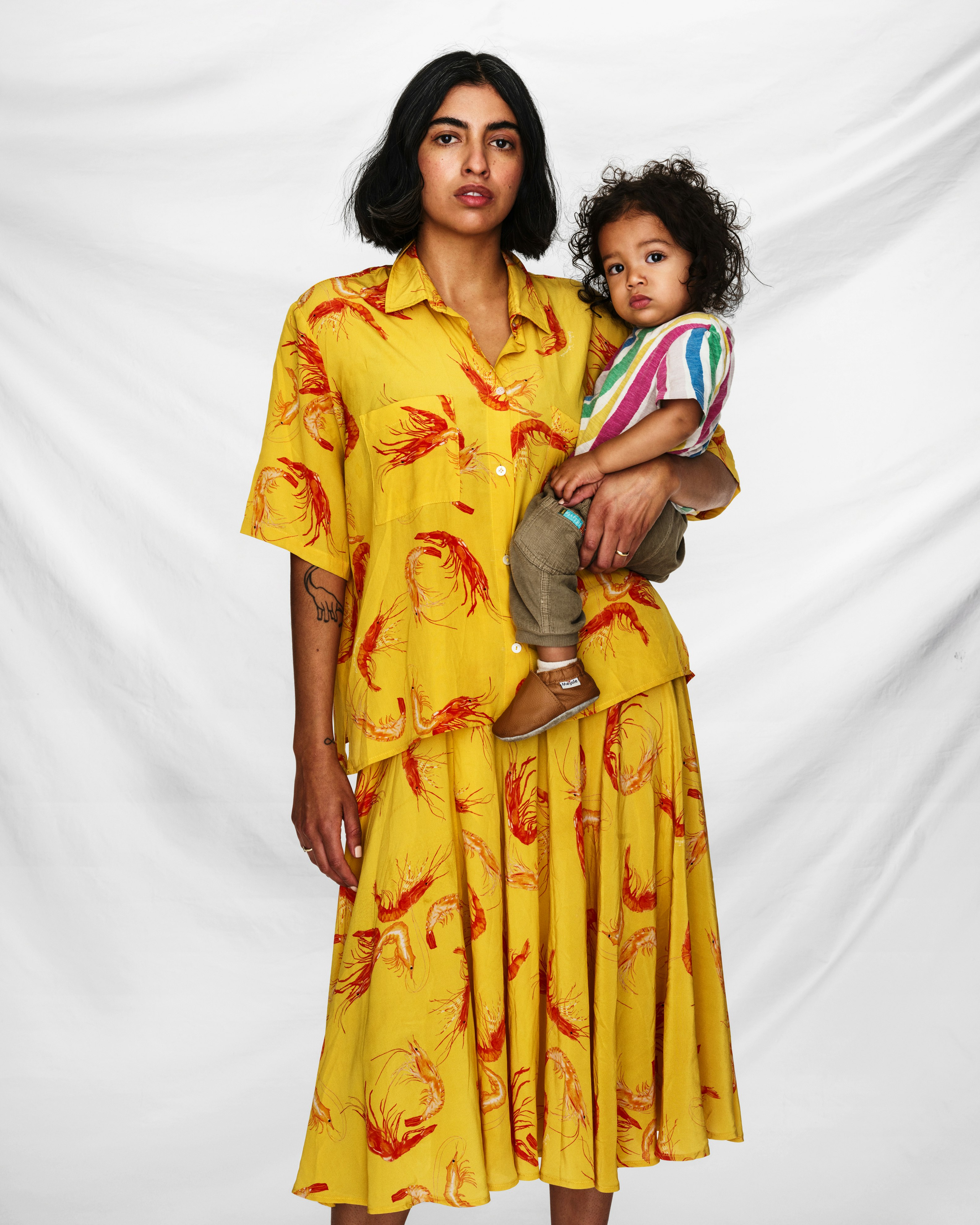 Person wearing a yellow dress with shrimp print, carrying a child, posed in front of a white cloth backdrop.