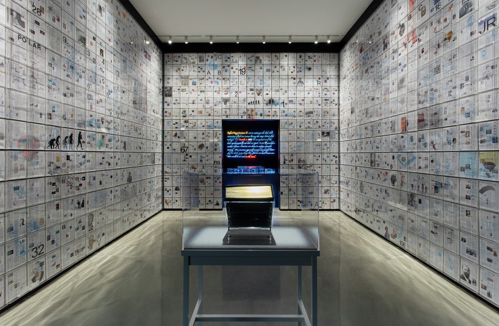 Art gallery with a thick encyclopedia volume encased in glass at the center of the room, with pages of the book lining the walls from floor to ceiling.