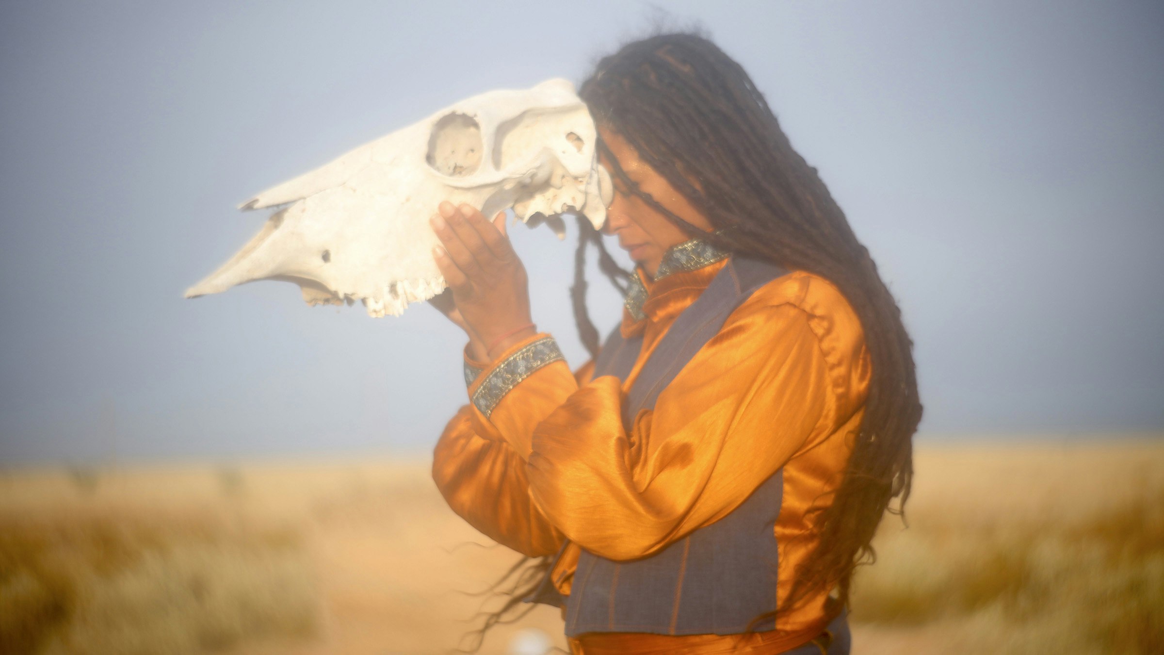 A photograph of a human figure with eyes closed wearing an orange jumpsuit holding a large white animal skull to her head.