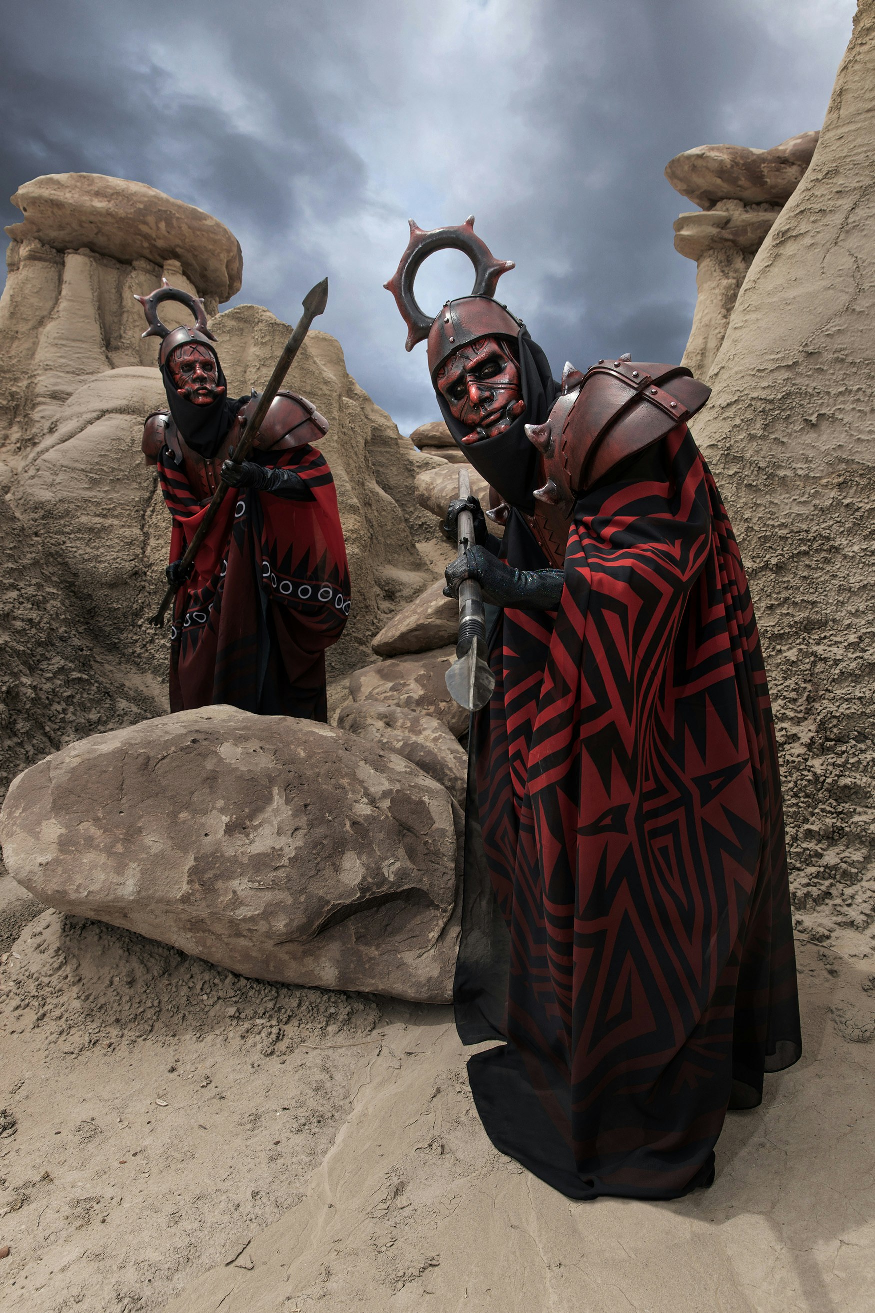 A photograph of two mean in red capes and bronze headdresses surrounded by large beige stones.