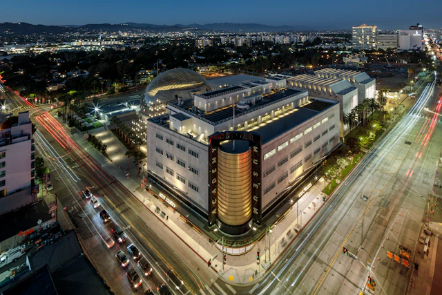 An aerial view photograph of a brightly lit building that takes up an entire city block. A gold column goes up the front in the center of the building.