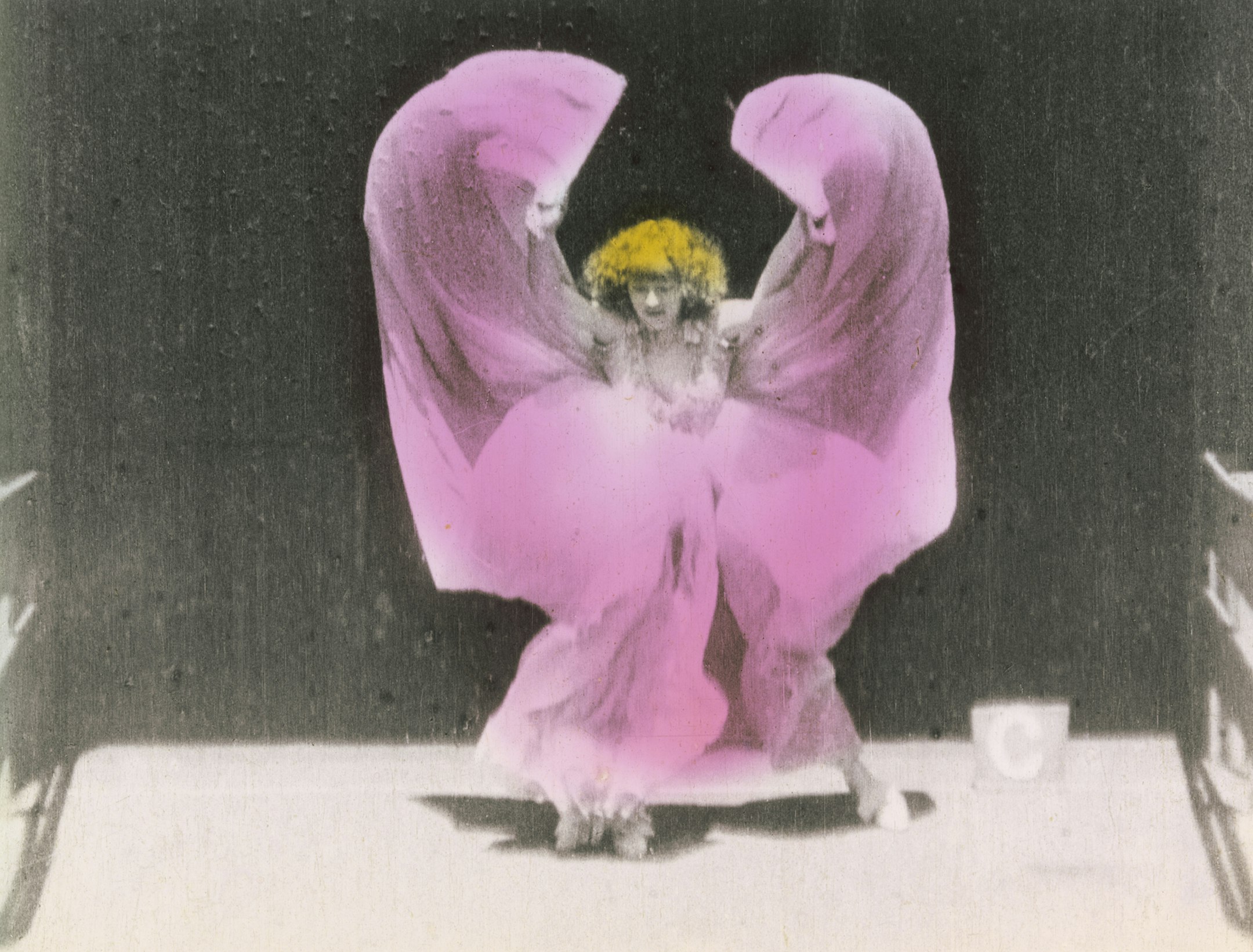 Hand-painted film still of woman in motion, dancing in a long, flowing skirt; the skirt is tinted pink and her hair is yellow.