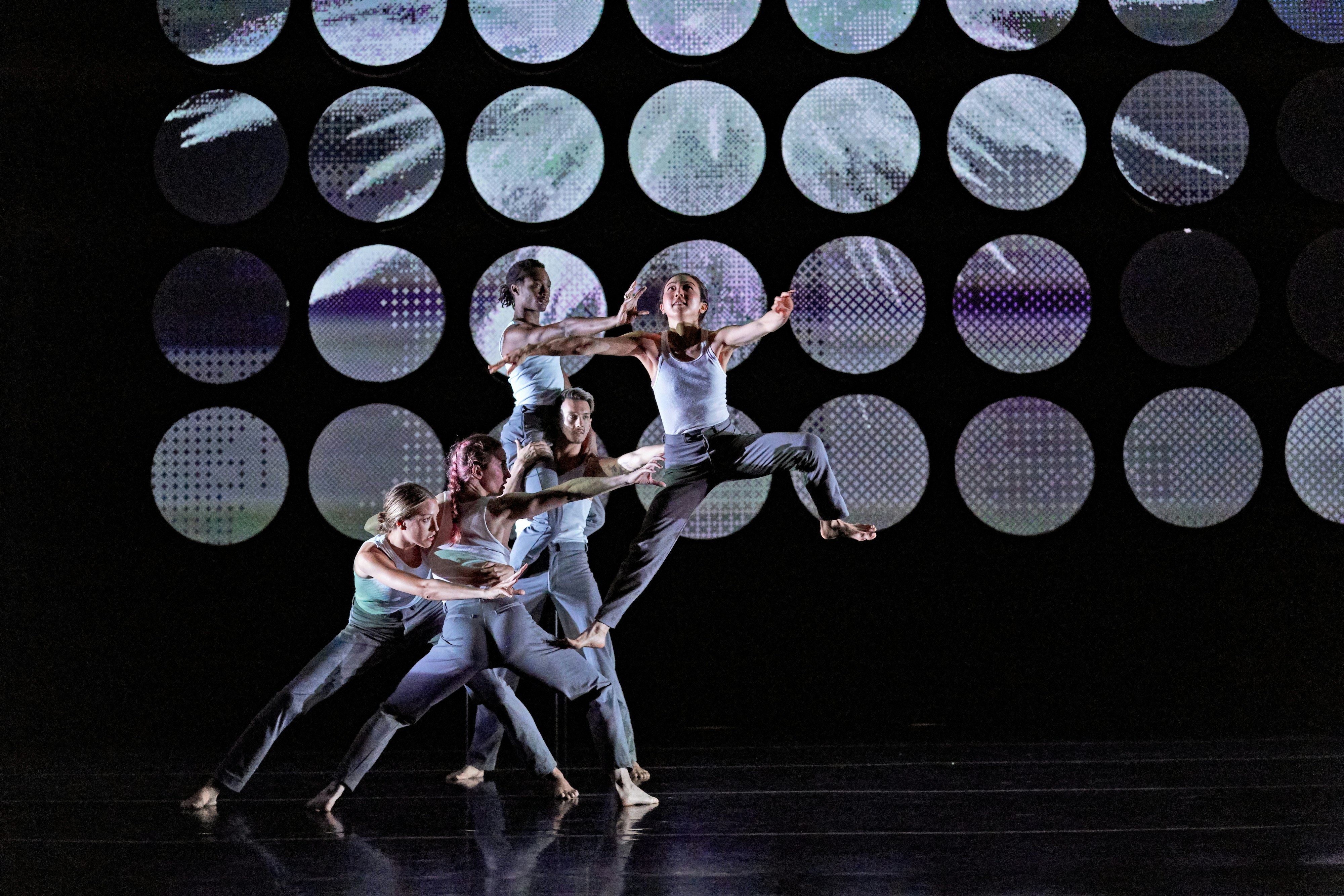 A group of dancers on stage, against a digital background of patterned circles. The dancers are in a formation going upward in space.