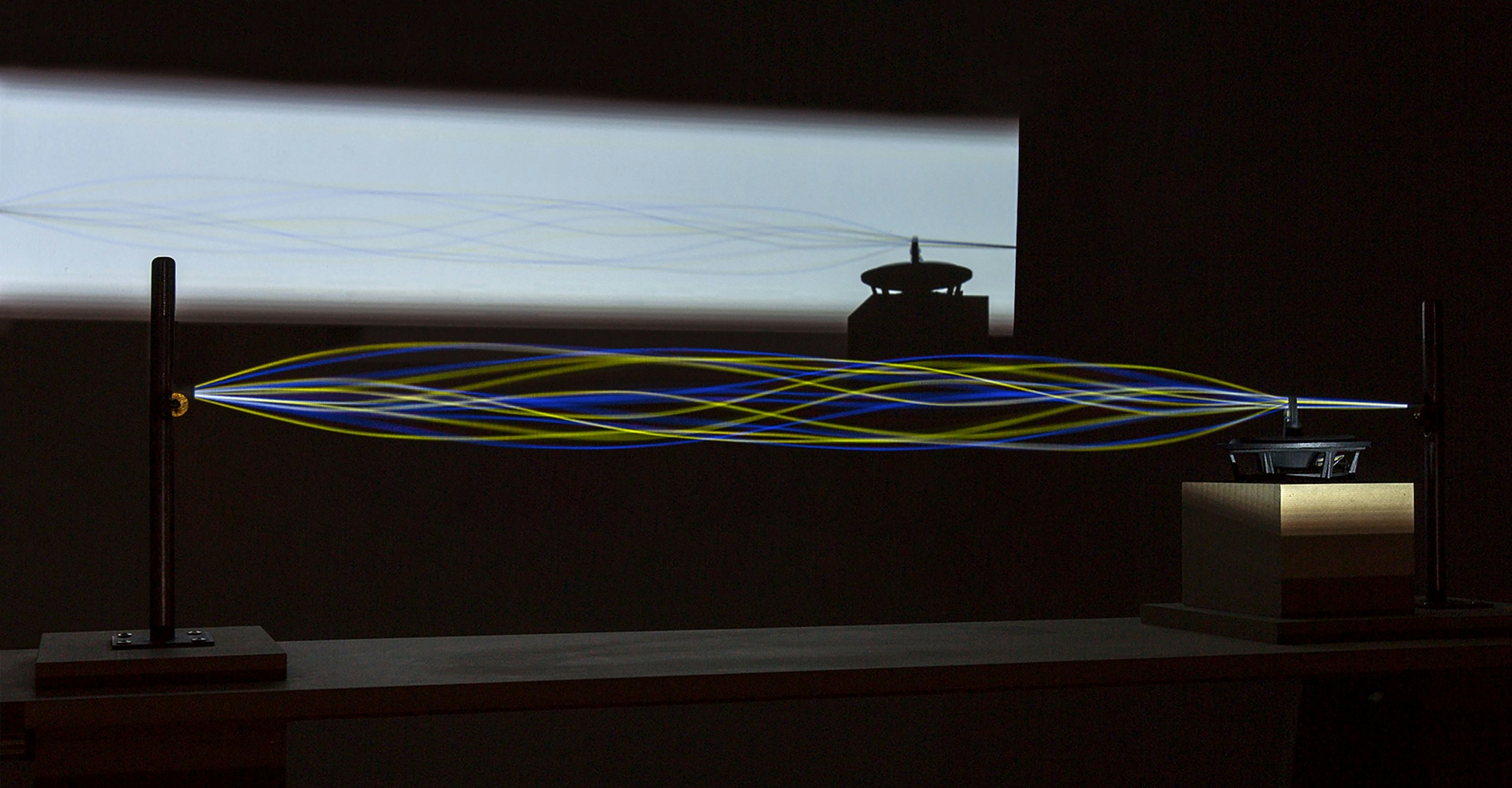 A string held between two mounted points, depicting a moving soundwave; its silhouette is projected onto a wall.