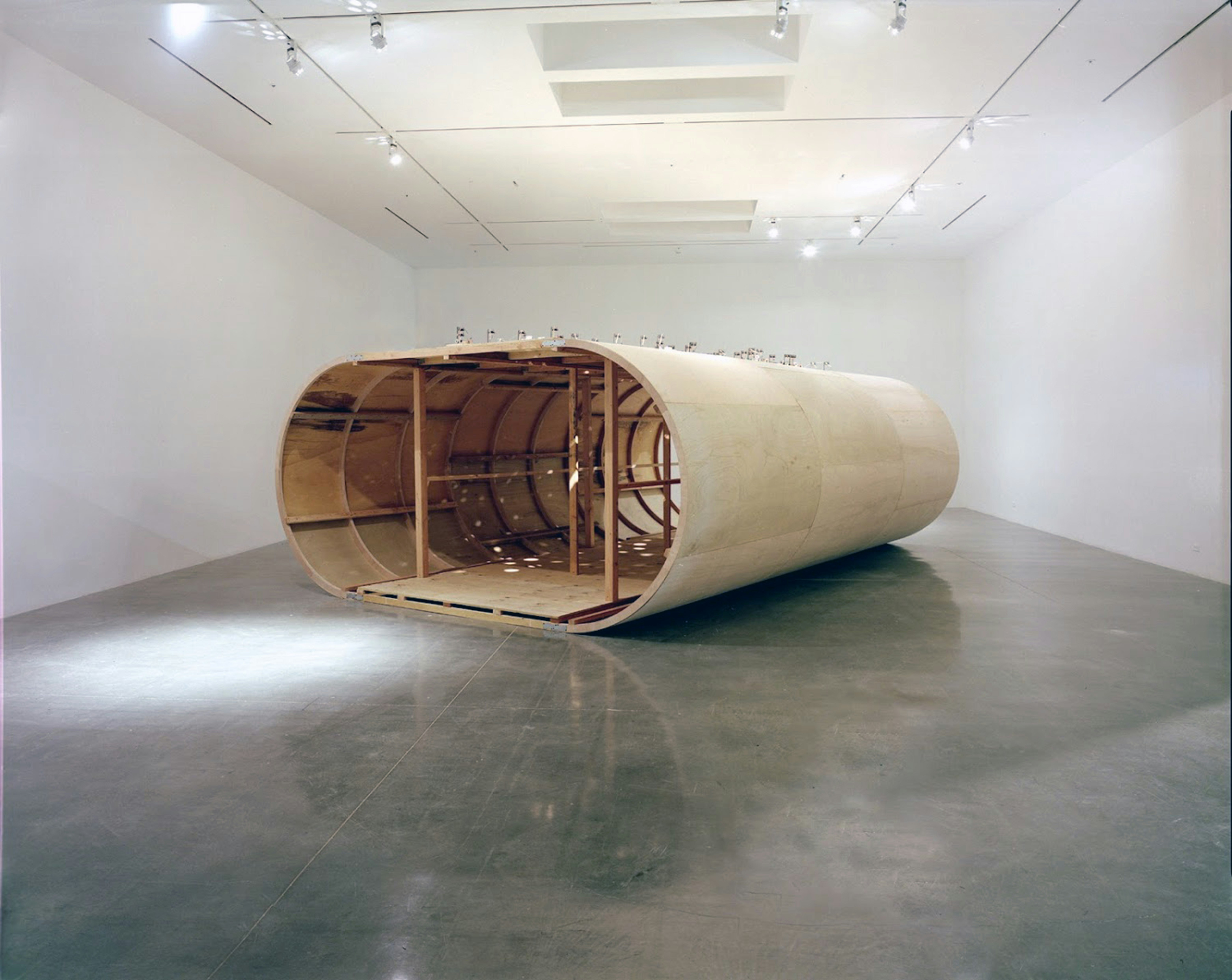 A photograph of a large oval wooden structure in a white walled room on a concrete floor.