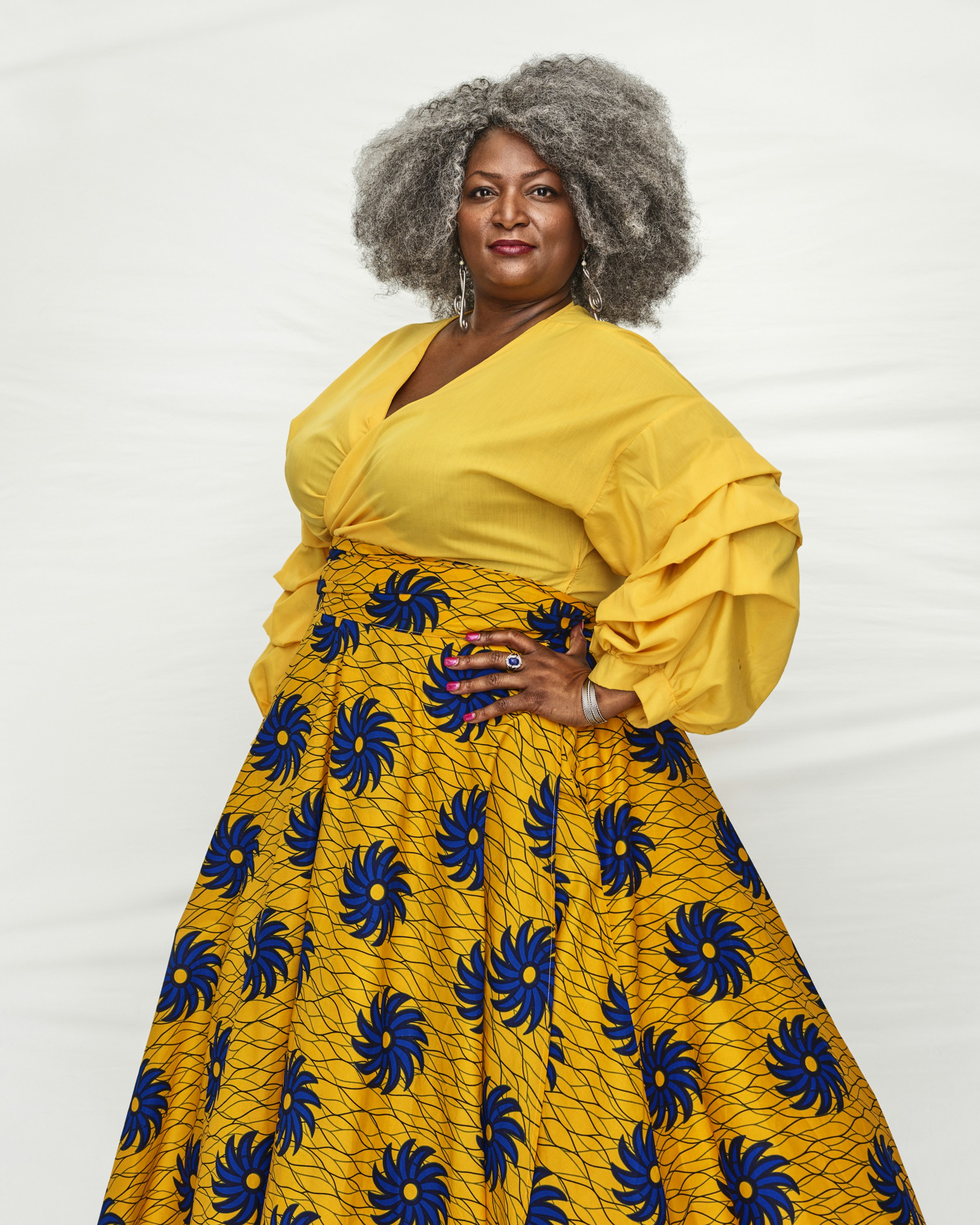 A black woman with grey curl hair stands with her hands on her hips. She's wearing a vibrant yellow shirt and a yellow skirt with bright blue flowers all over it.