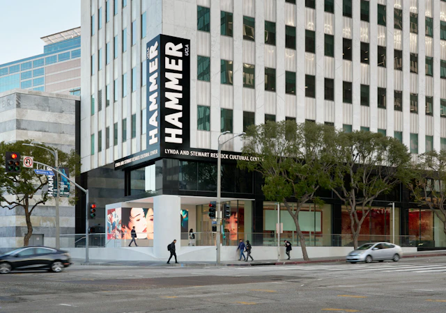 Exterior view of building with signage that reads: “Hammer UCLA” “Lynda and Stewart Resnick Cultural Center.” Moving cars trail across the main street, with museum banners on street poles.