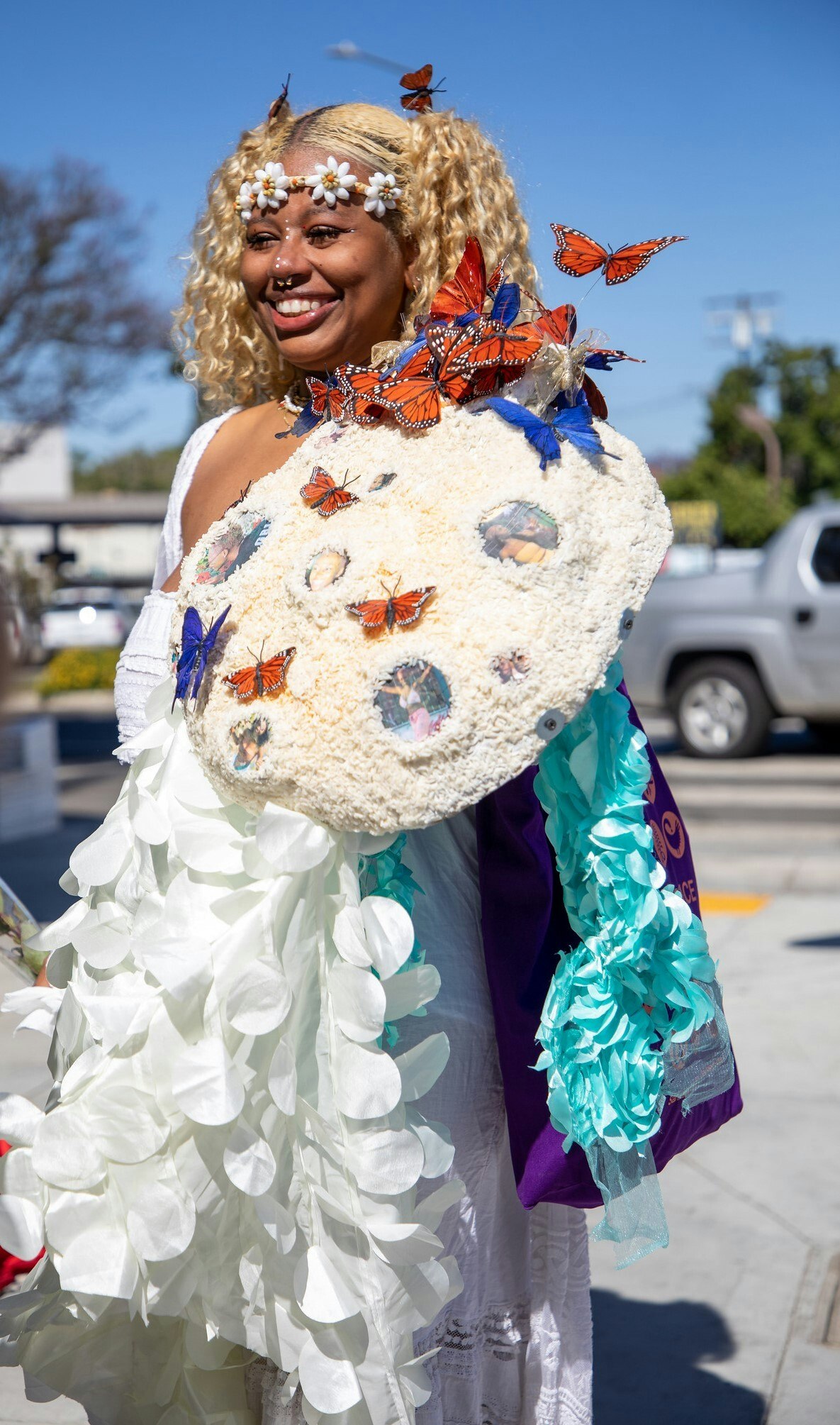 A photograph of a smiling woman with blonde hair. She's wearing a daisy chain headband, tortoiseshell butterflies are in her hair and on her white dress.