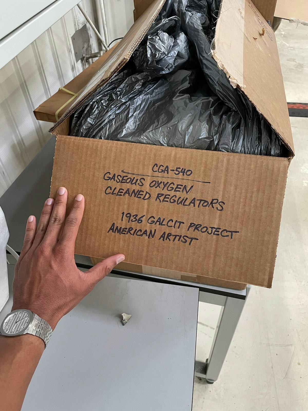 A photograph of a cardboard box with a silver garbage bag in it. A human hand reaches for it.