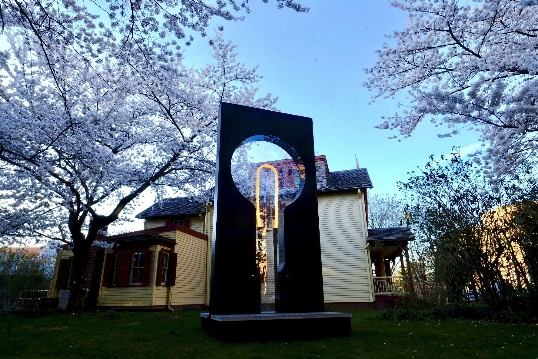 Large, flat sculpture with the outline of a bulb and a lit filament in the center, in front of a two-story house and surrounded by cherry blossom trees.