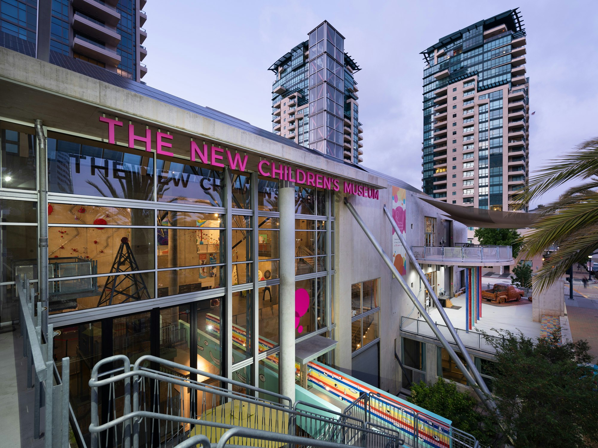 Exterior view of a building with a glass facade and a colorful bridge, with signage that reads: “The New Children’s Museum” in pink.