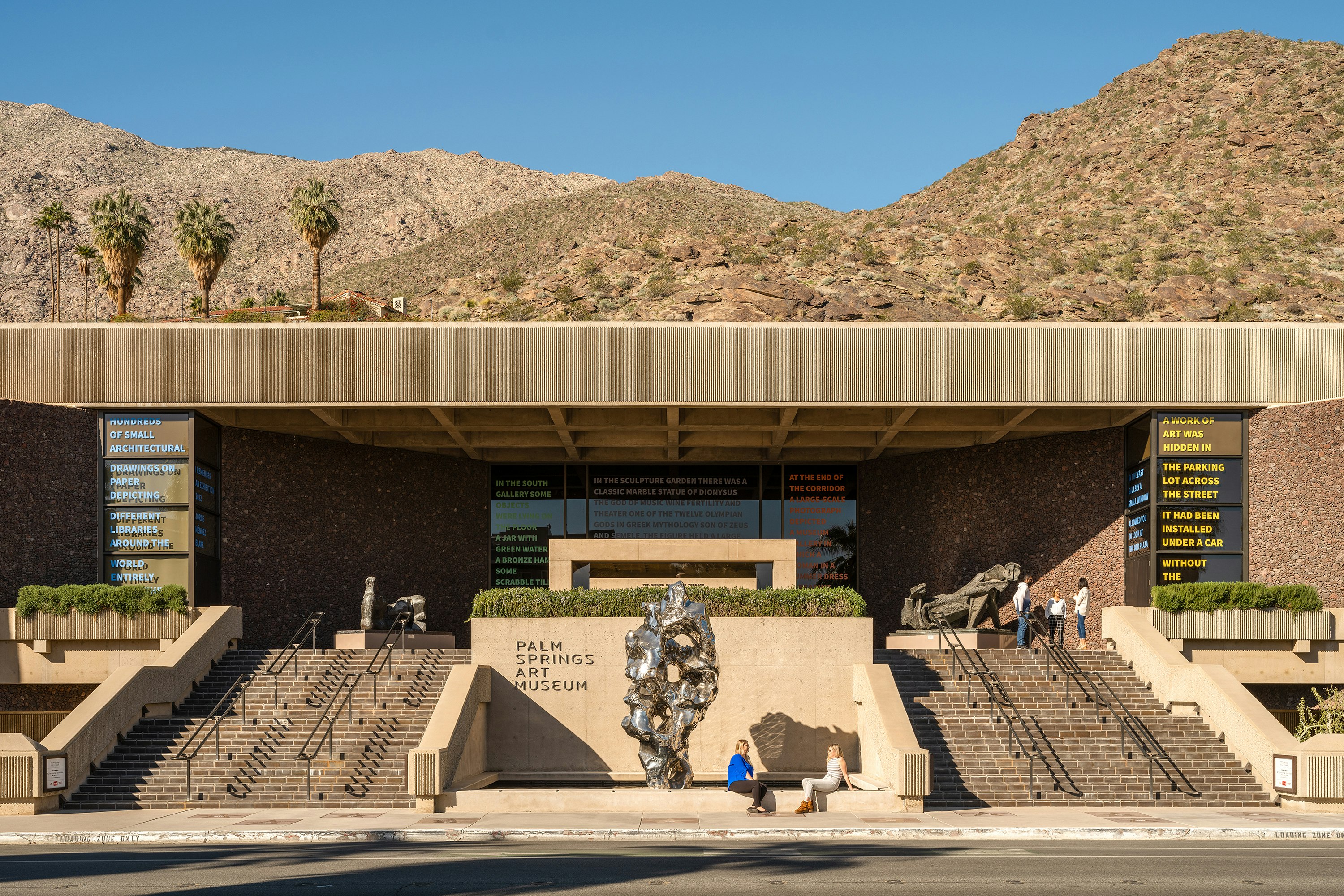 A photograph of a beige-colored, low, mid-century modern building with steps leading up to the front. Brown desert hills rise behind it.