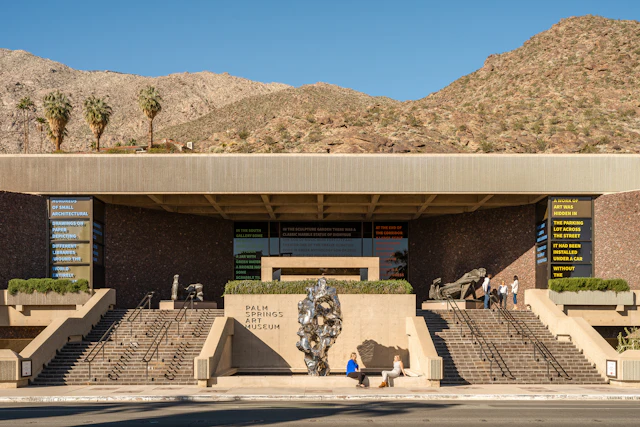 A photograph of a beige-colored, low, mid-century modern building with steps leading up to the front. Brown desert hills rise behind it.