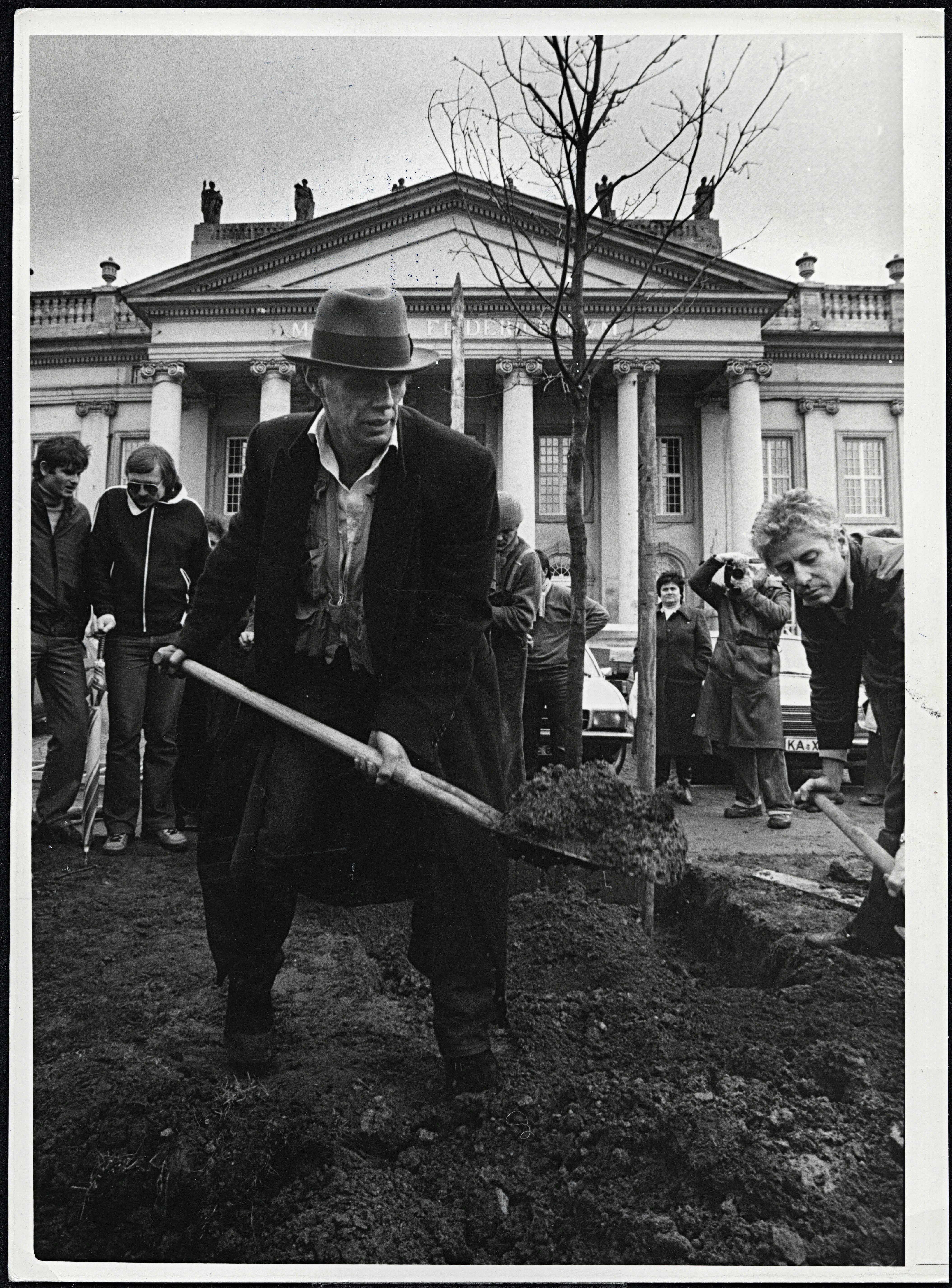 Man wearing long coat and hat digging hole to plant oak tree in front of museum building.
