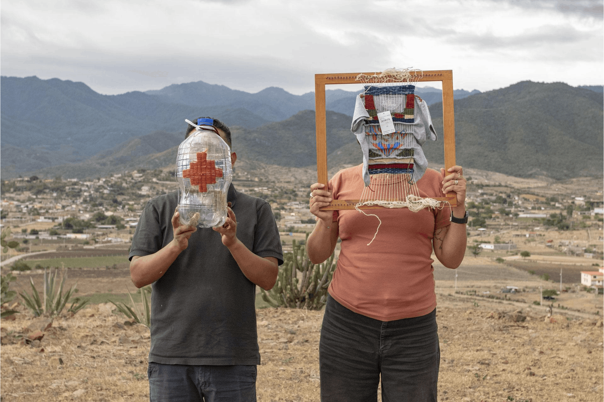 Color photograph of two people standing in front of a desert landscape. The figure on the right holds a wood frame with weaving in front of their face. The figure on the left holds a plastic water bottle in front of their face.