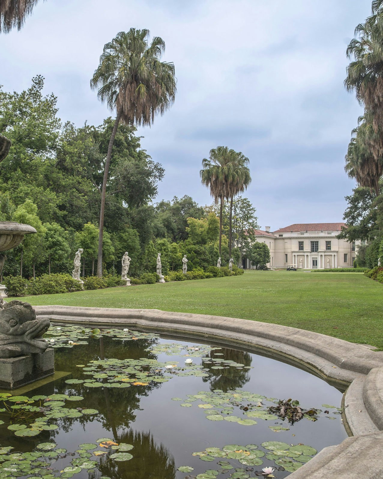 A photograph of a stone fountain with lily pads in the water. A green lawn vista flanked by palm trees and classical stone statues recede to a cream stone building with an orange tiled roof.