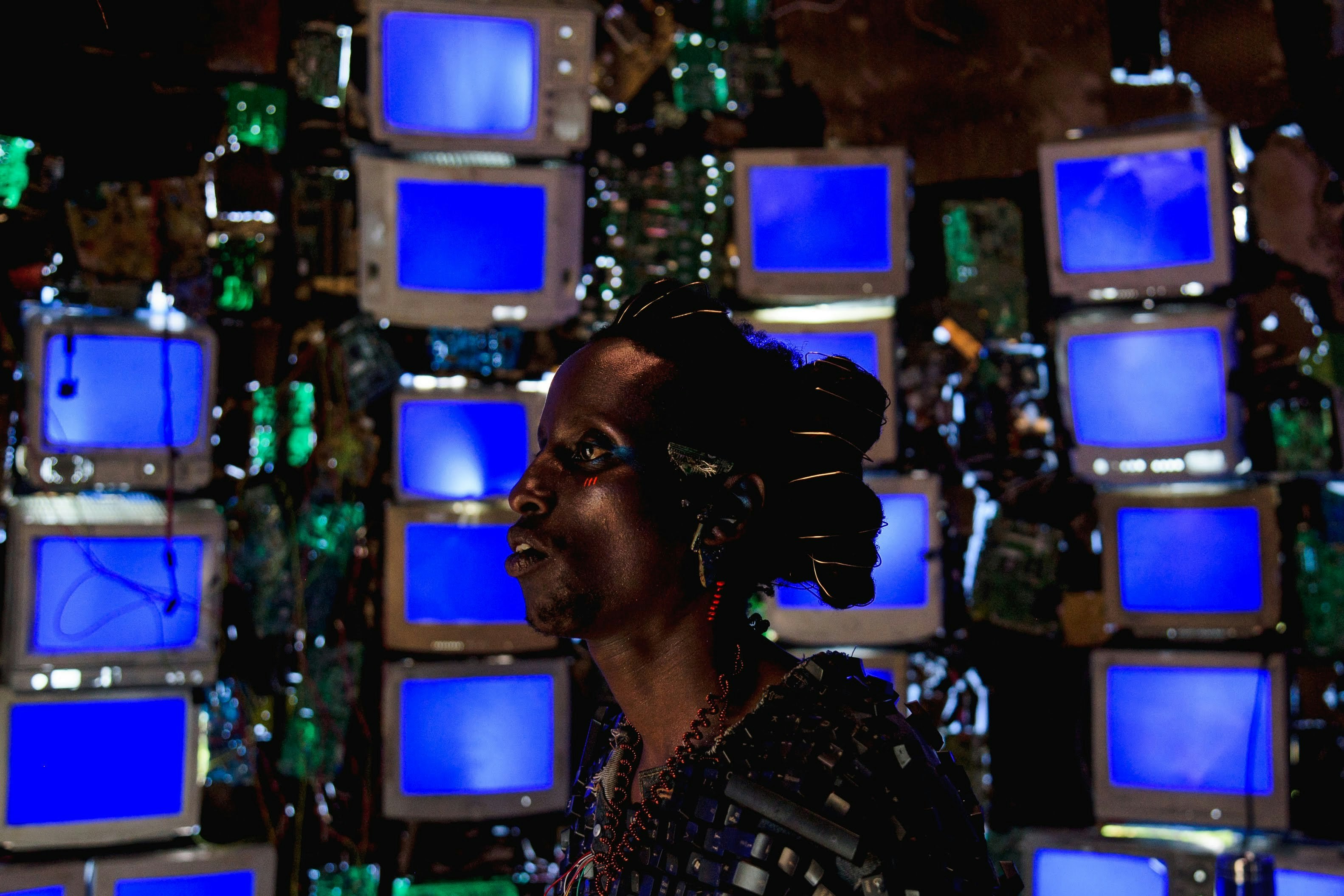Person with hair and ear accessories in front of 4 columns of television sets with blue screens.